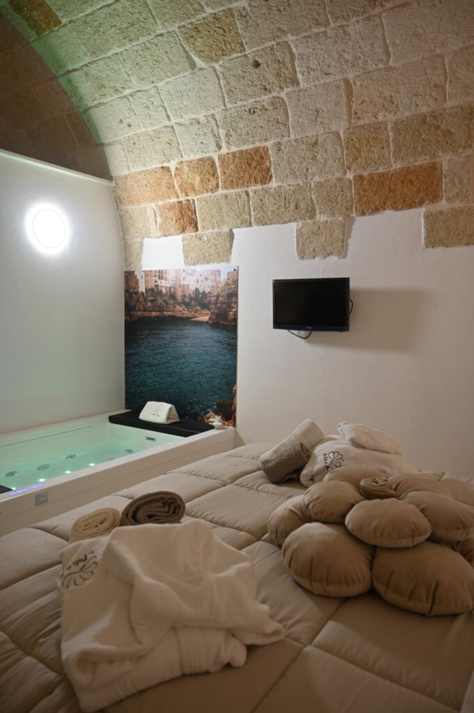 Camera con letto e idromassaggio accanto, una gigantografia di polignano è dietro al poggiatesta dell'idromassaggio a led, il muro in pietra di tufo con la volta a botte si inarca e rende l'ambiente confortevole, la luce soffusa e le coperte in tinta con la pietra rendono la stanza accogliente.