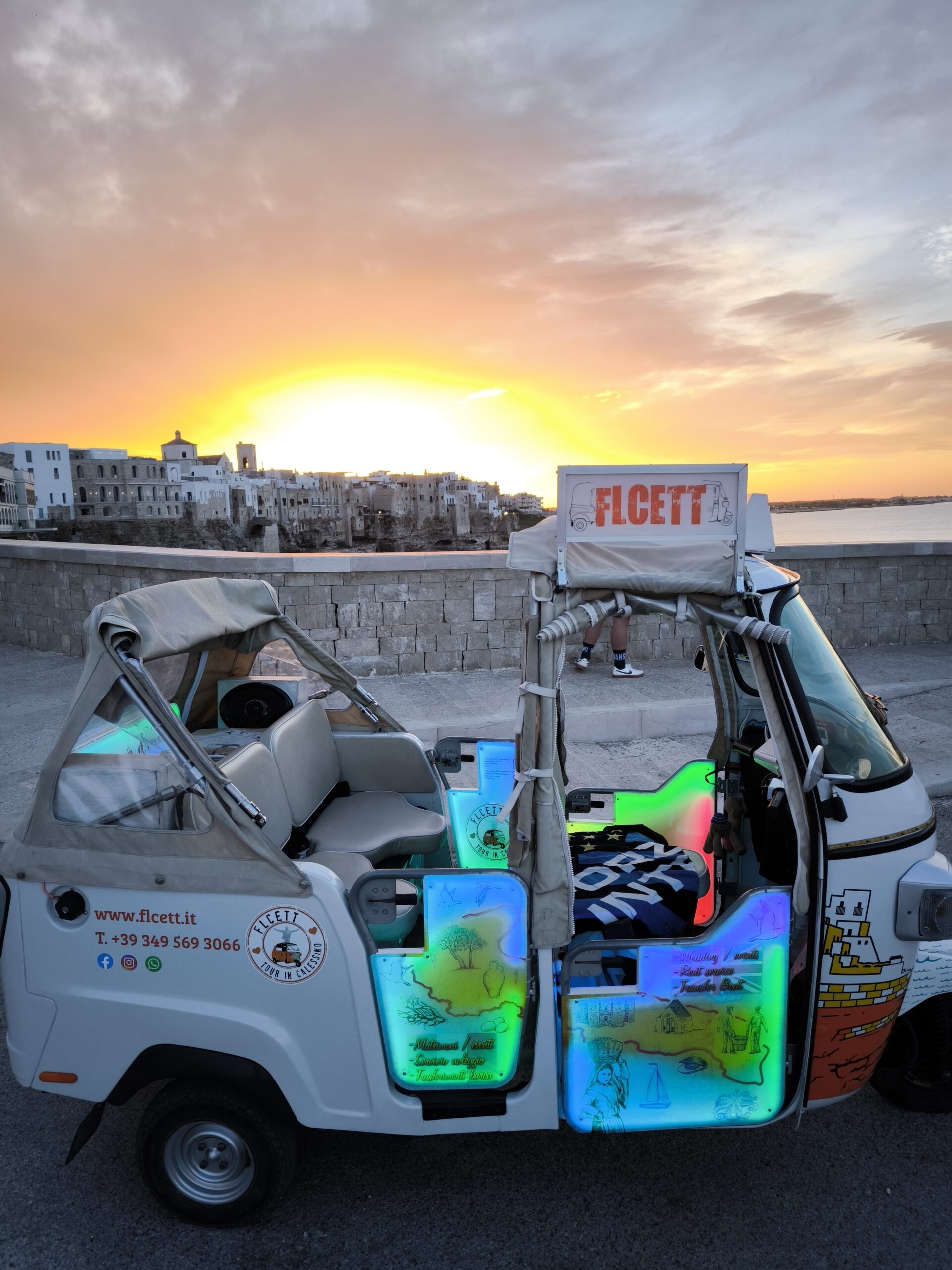 Ape Calessino turistico decorato, fermo sul lungomare di Polignano a Mare al tramonto, con vista panoramica sul centro storico e il mare Adriatico.